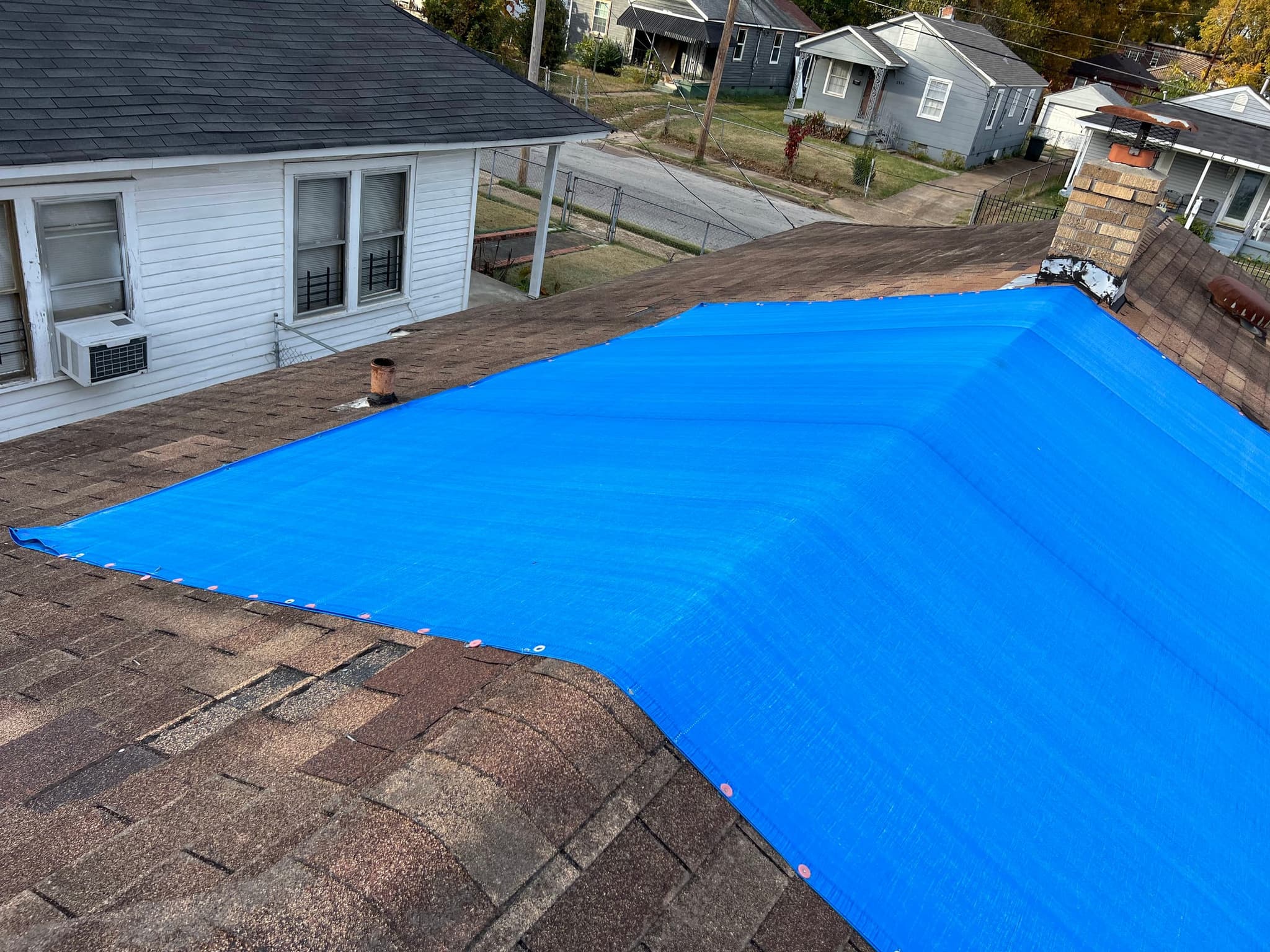 Blue tarp covering part of a shingle roof on a white house, photographed from the roof with neighbouring houses below