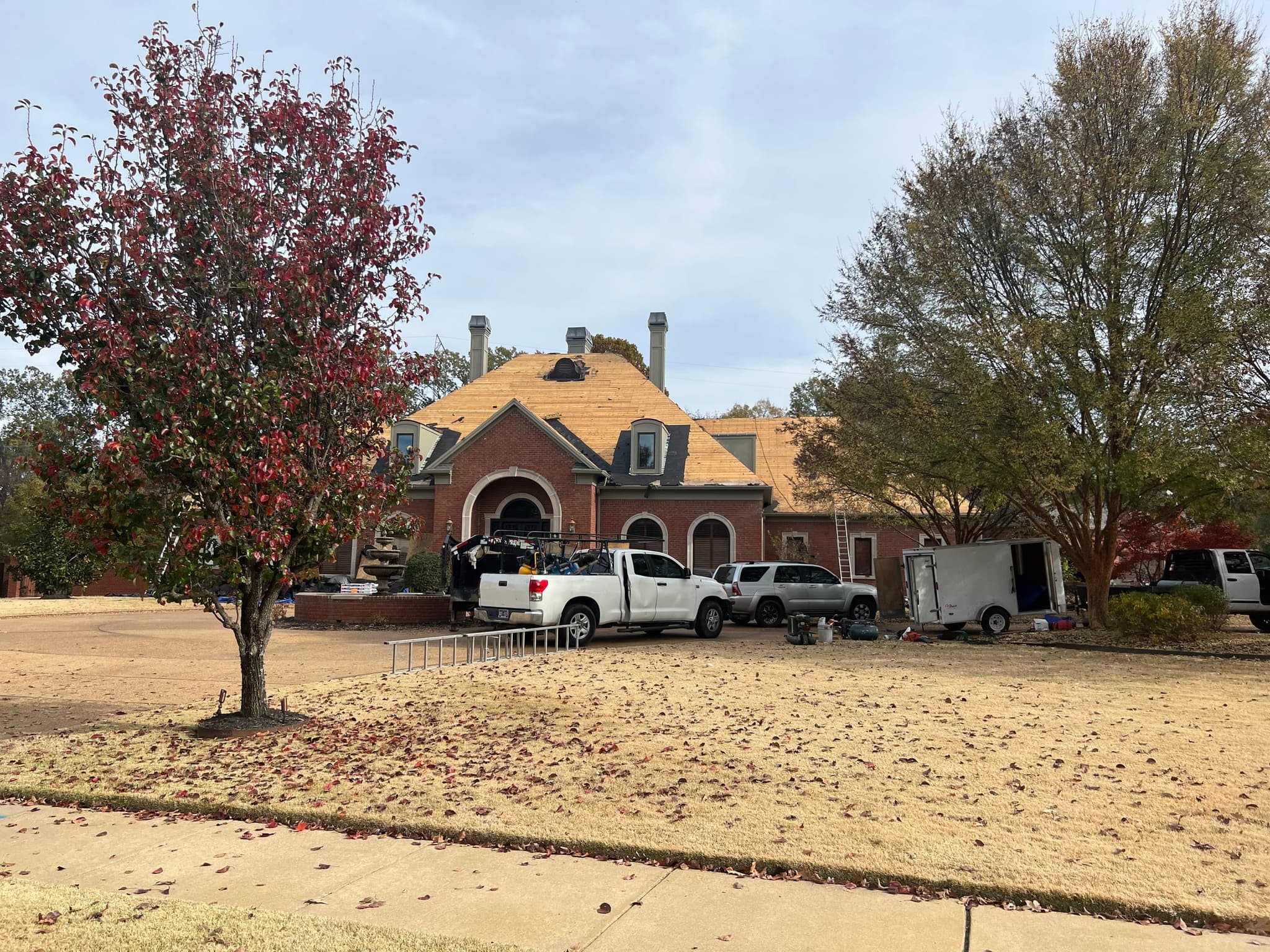 Large brick house with an arched entry whose roof is stripped to bare decking, with crew trucks and a trailer parked in front