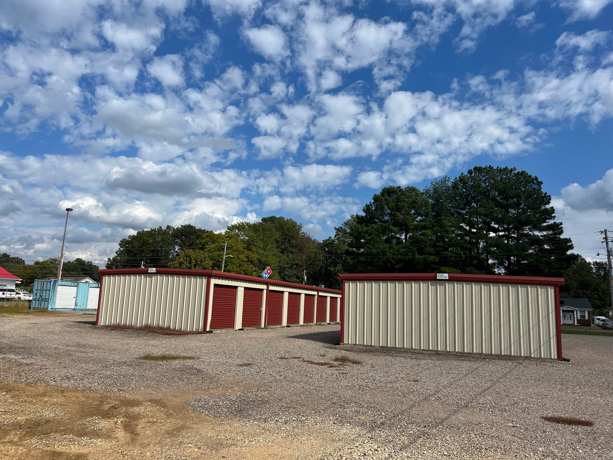 Rows of metal self-storage buildings with red roll-up doors and cream walls on a gravel lot under a blue sky with scattered clouds