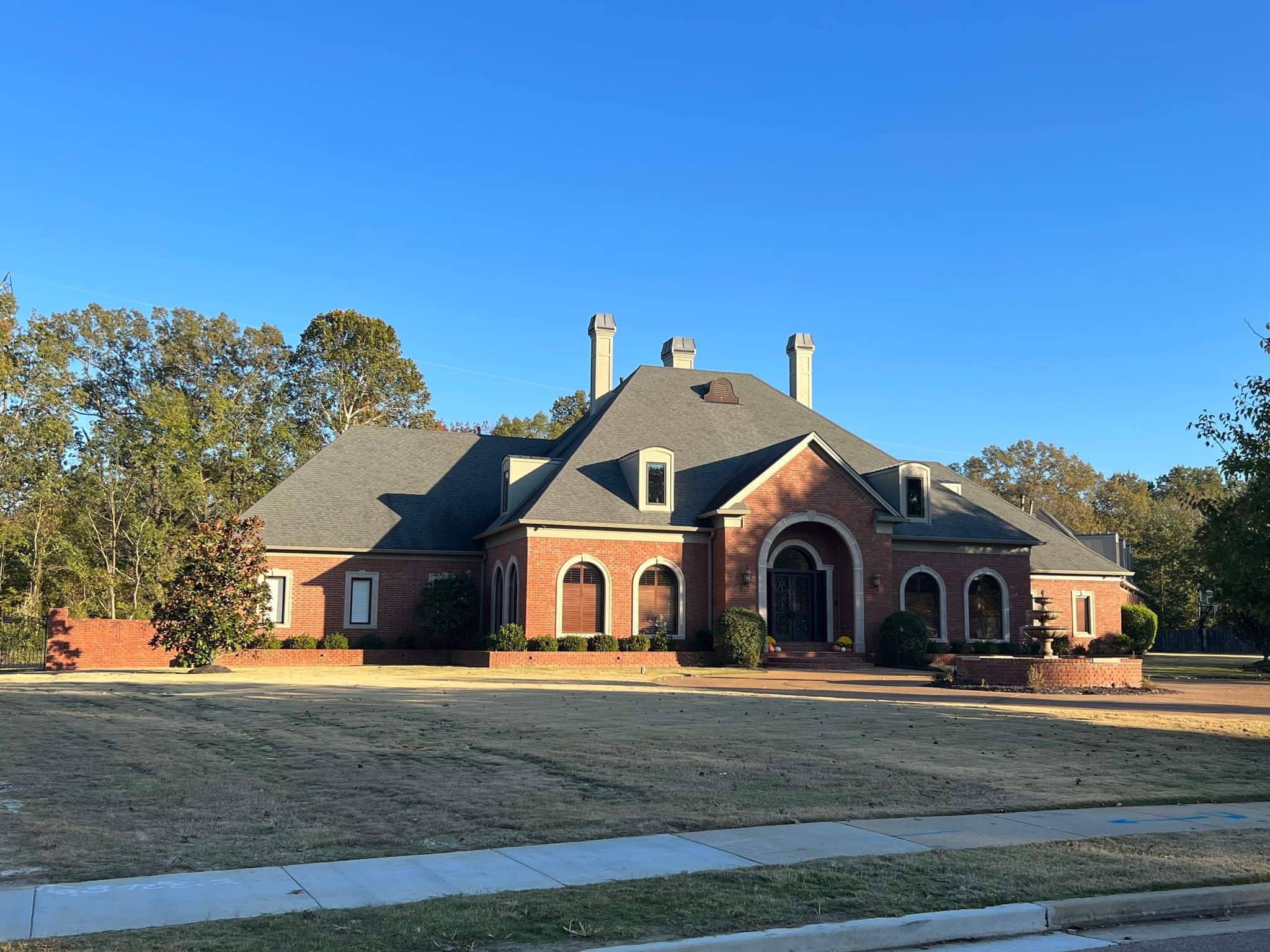 Wide view of a large red brick home with arched windows and a courtyard fountain, existing grey shingle roof before replacement