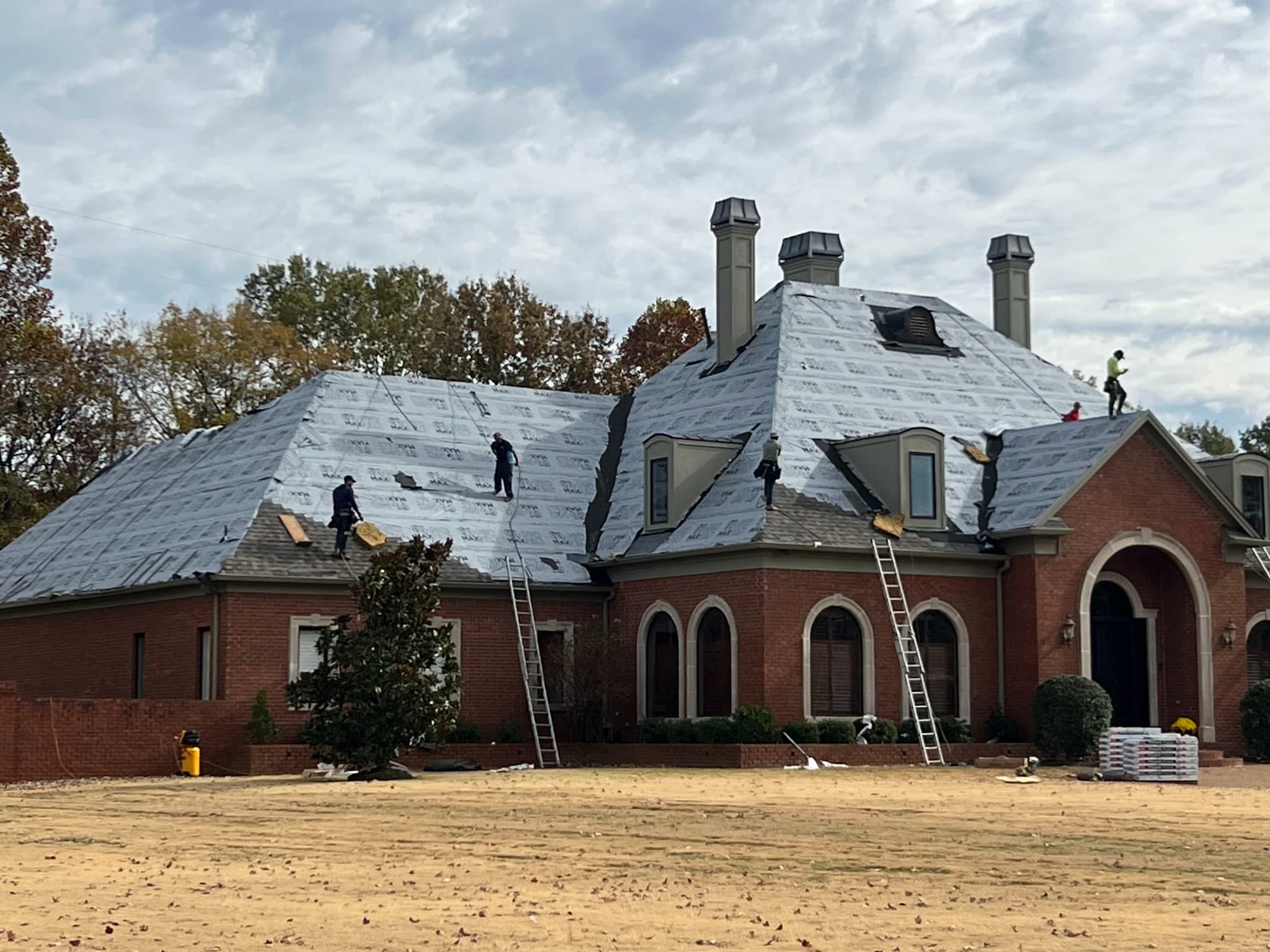 Roofing crew installing synthetic underlayment on the stripped roof deck of a large brick home, with old shingles still on one section