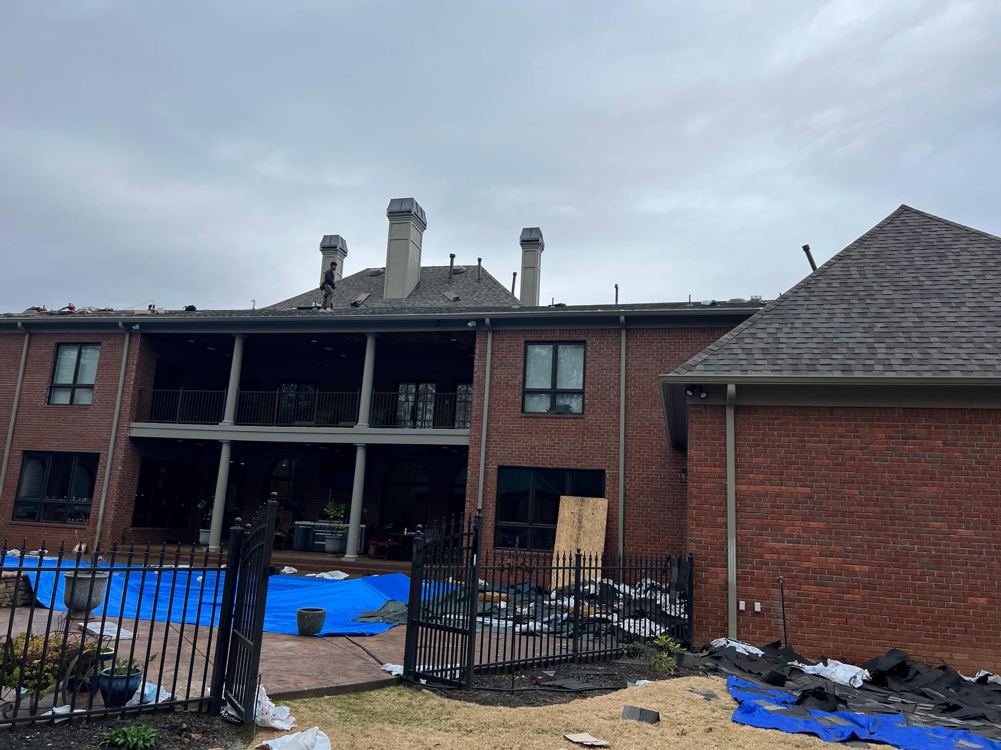 Rear of a large brick home with a covered balcony; the pool and patio are covered with blue tarps while a roofer works above