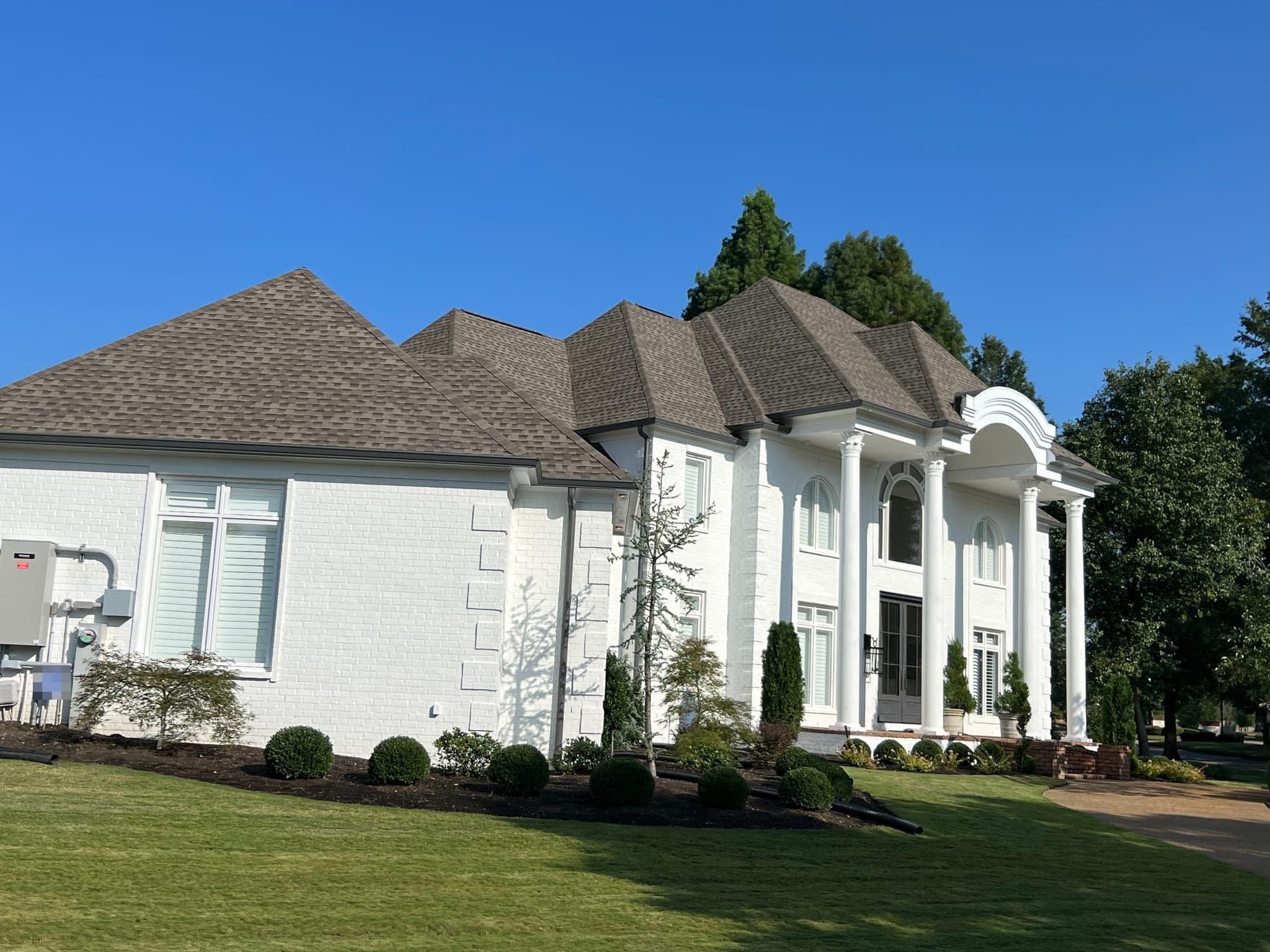 Large white-painted brick home with a columned portico and a warm grey architectural shingle roof under a clear blue sky