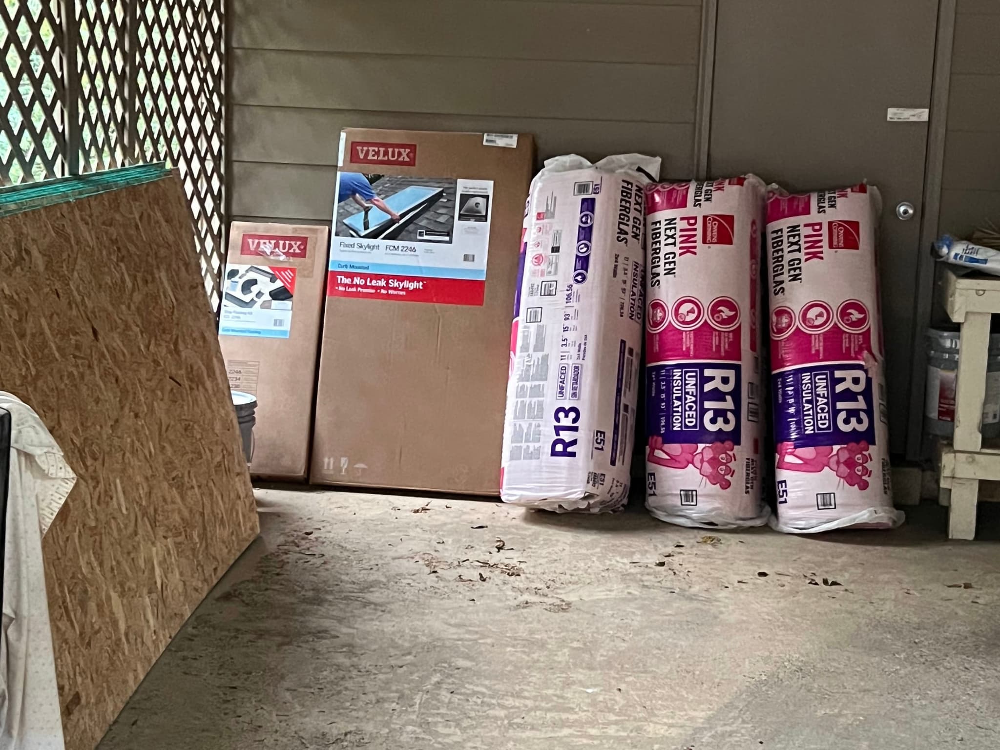 Materials staged against a carport wall: Velux skylight boxes, pink R13 fibreglass insulation and sheets of OSB decking